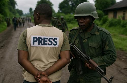 Un soldat armé contrôle un journaliste sur une route boueuse entourée de végétation. L’homme de presse, reconnaissable à sa veste marquée "PRESS", se tient calmement les mains derrière le dos